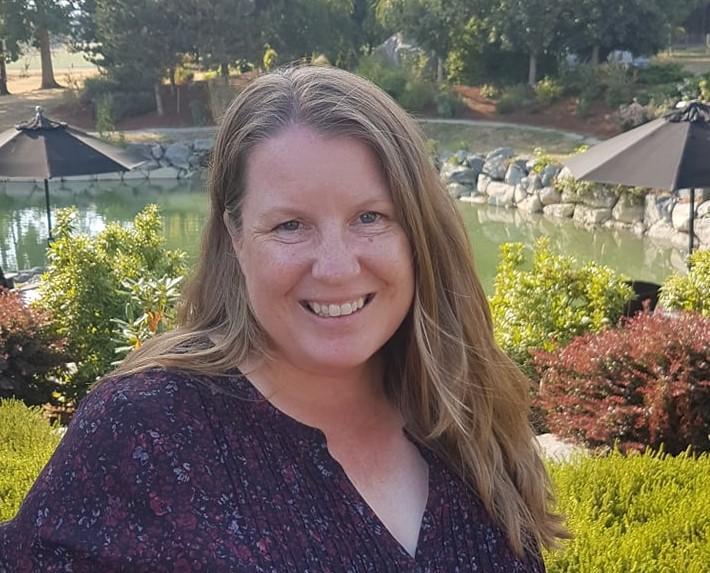 A woman stands in front a pond and garden smiling face-on into the camera.