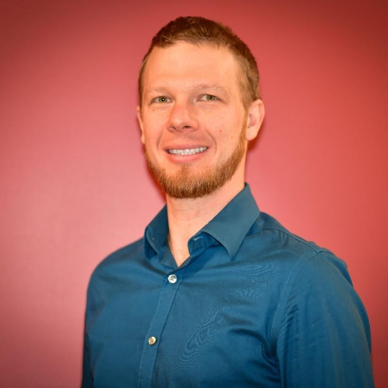 photo of a light-haired main with a beard wearing a denim blue shirt standing in front of a dark pink background