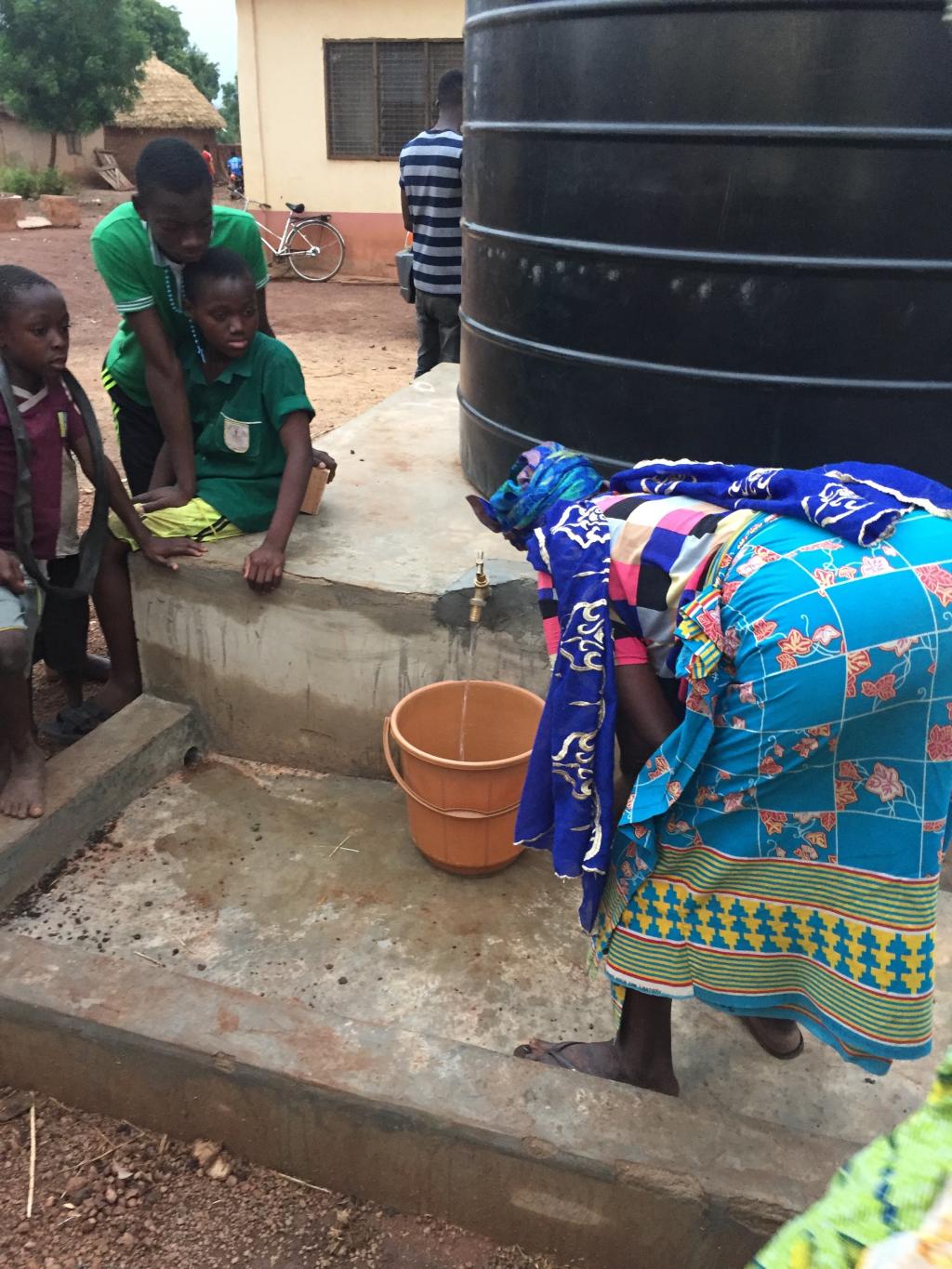 person bends down to place a bucket under a water spigot attached to a large tank as children watch