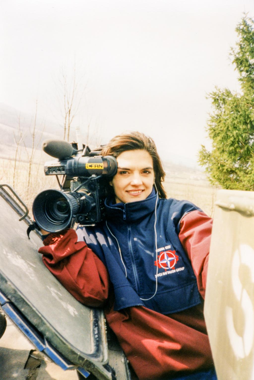 A woman smiles at the camera while holding a television camera on her right shoulder.