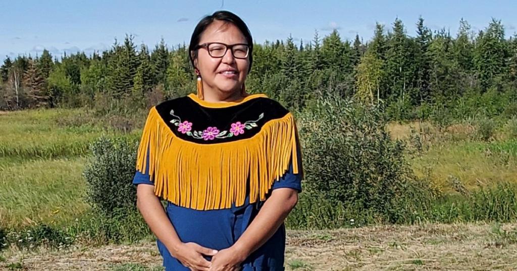 Doreen Arrowmaker poses for a photo. She stands with a treeline behind her and an embroidered print on her dress.