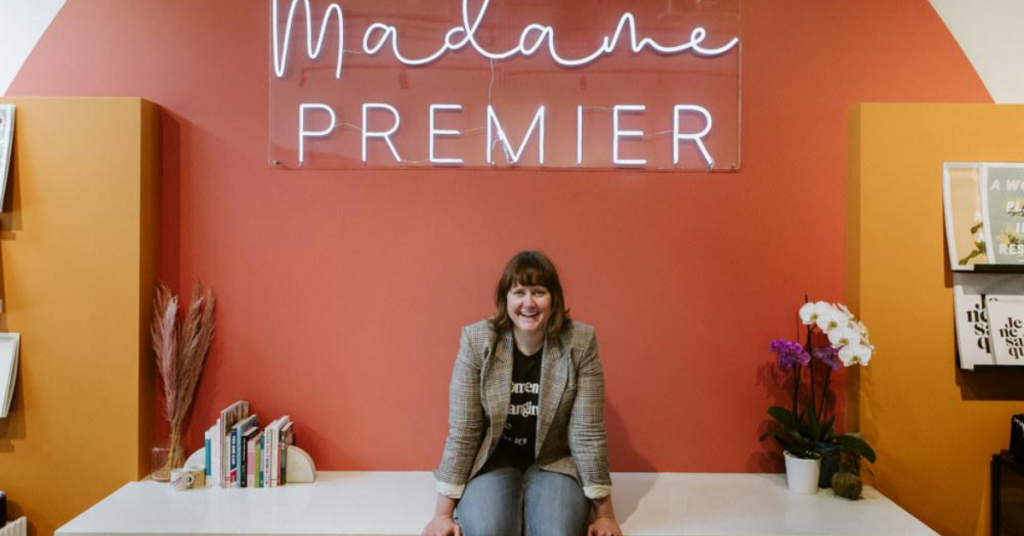 A woman sits on a table while smiling. The words, Madame Premier, can be seen on the wall behind her.