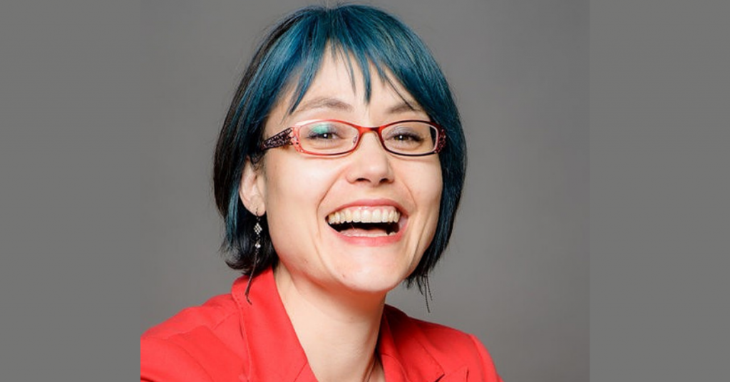 A woman with blue hair and glasses smiles for a head shot.