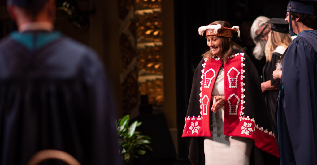 A woman in a button blanket crosses the stage at an RRU convocation ceremony.