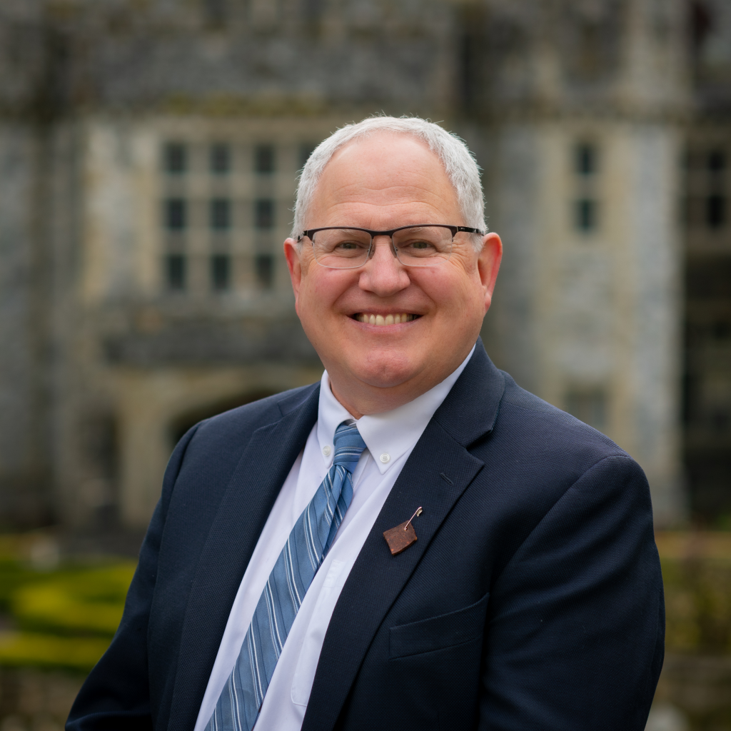 A headshot of Kevin Brewster standing outside Hatley Castle. He is wearing a suit and a Moosehide Campaign leather pin.