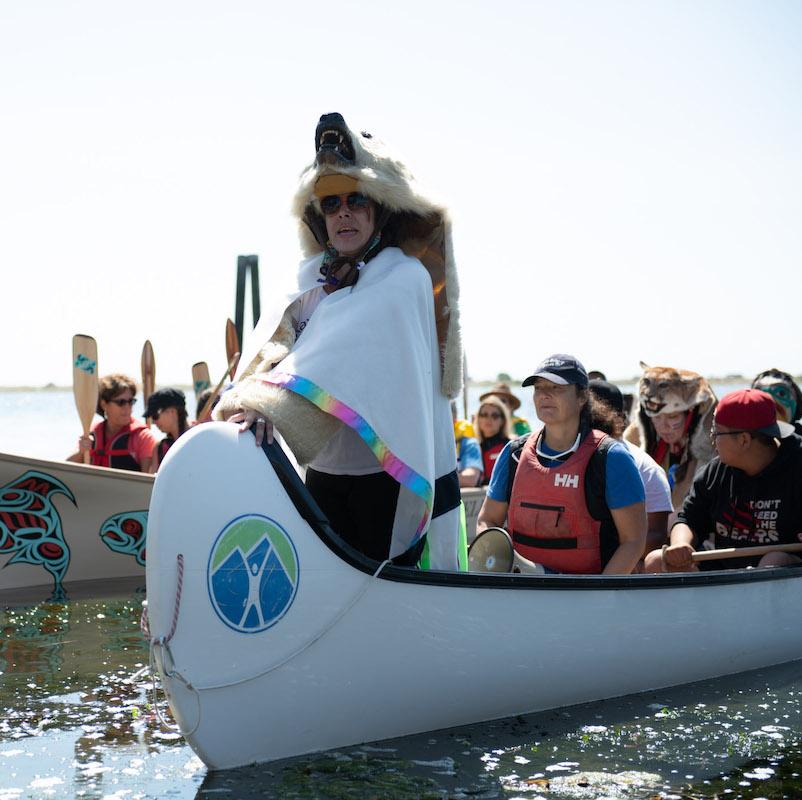 A woman wearing a bear skin in the font of a canoe requests permission to land.