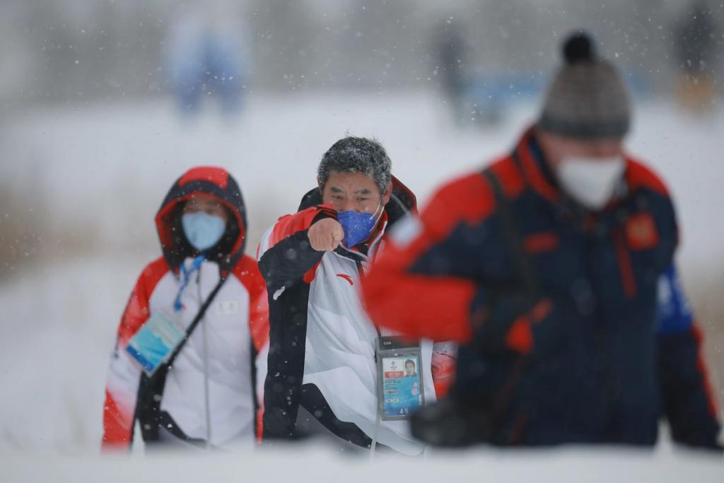 A masked man gestures forward while walking in the snow. 