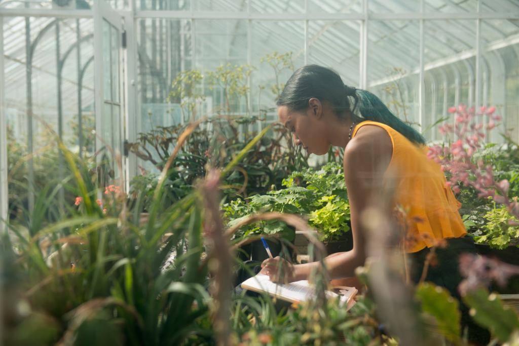 A woman takes notes surrounded by plants in a greenhouse.