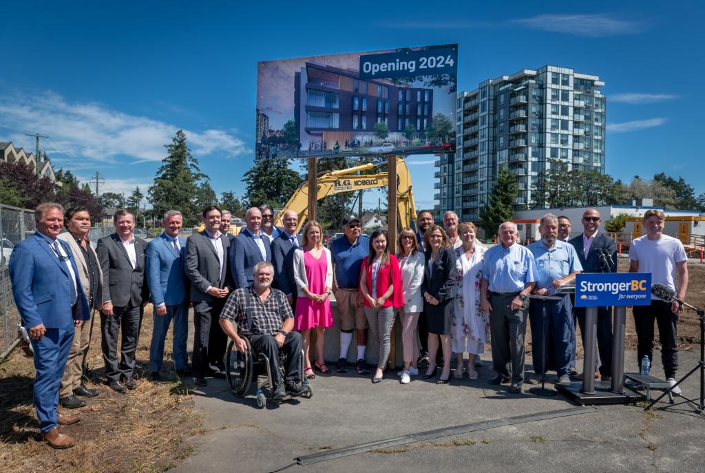 Dignitaries and partners stand together on the future grounds of the campus in front of a sign that says 'Opening 2024" with a rendering of the first building.