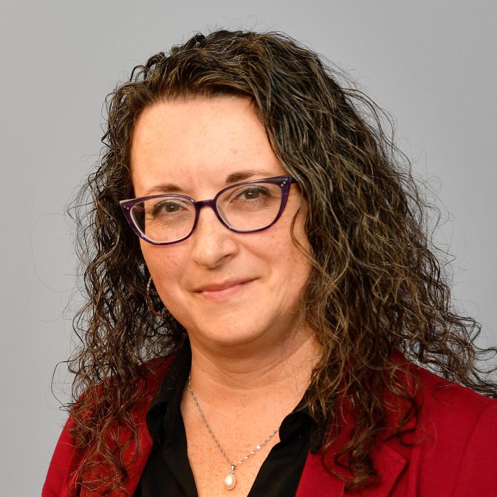 A woman poses for a headshot against a grey background. She looks happy.