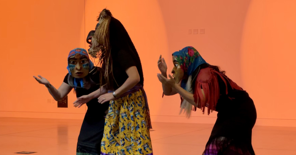 ANSWER2 Drum Group performs. Three masked figures hunch in the foreground with a wash of orange light behind them.