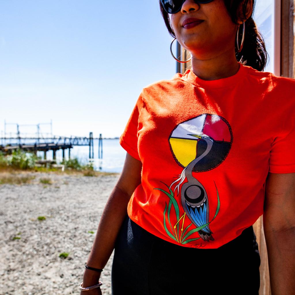 A person leans against the Boathouse wall at the Esquimalt Lagoon wearing an orange shirt with heron artwork by Butch Dick.