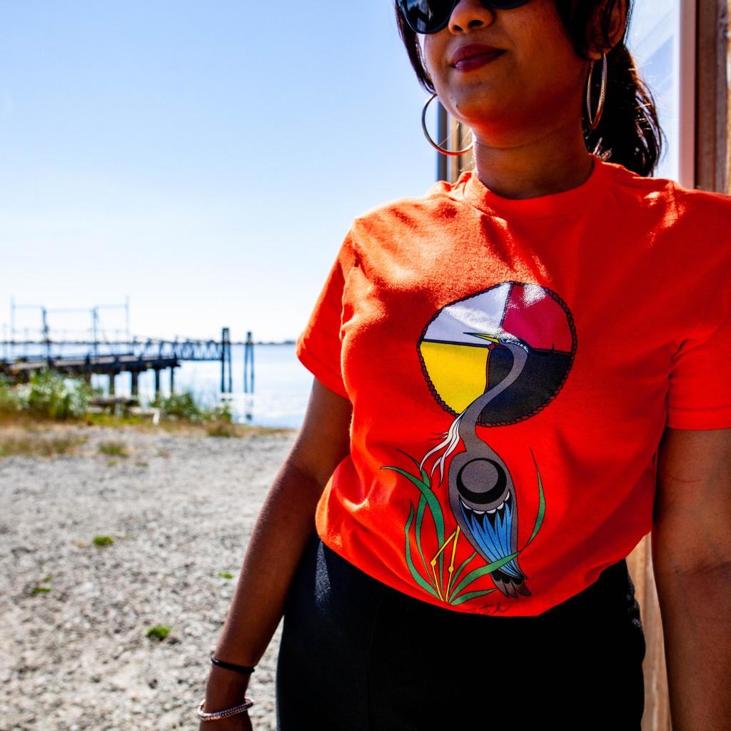 A person leans against the Boathouse wall at the Esquimalt Lagoon wearing an orange shirt with heron artwork by Butch Dick.  