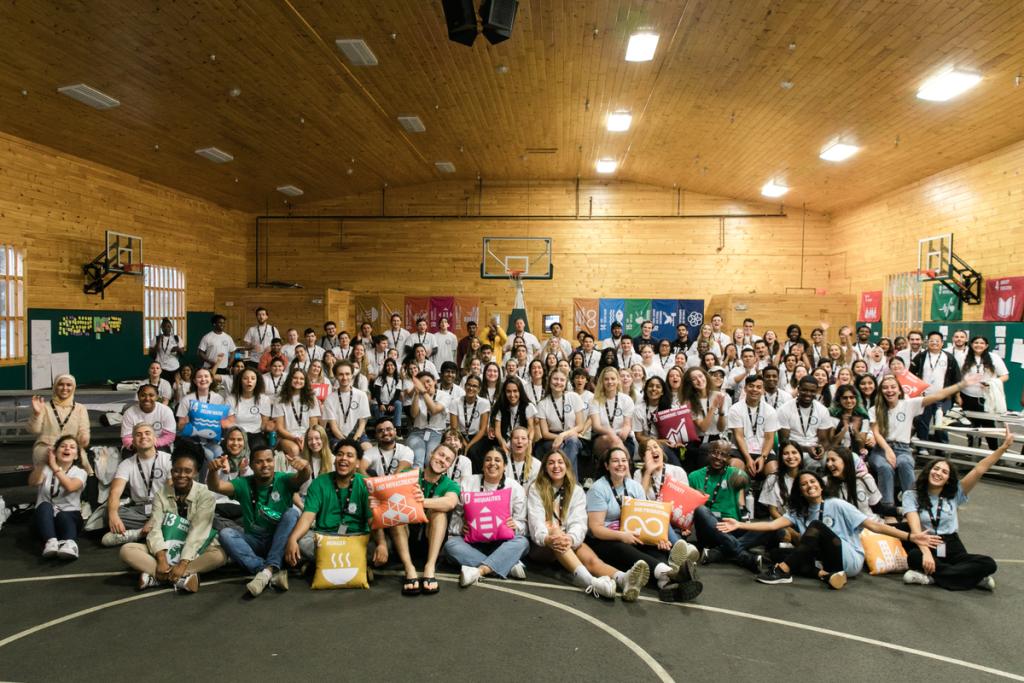 A group of young people sitting in a gymnasium wearing matching CAMP shirts and holding pillows with UN Sustainable Development Goals printed on them.