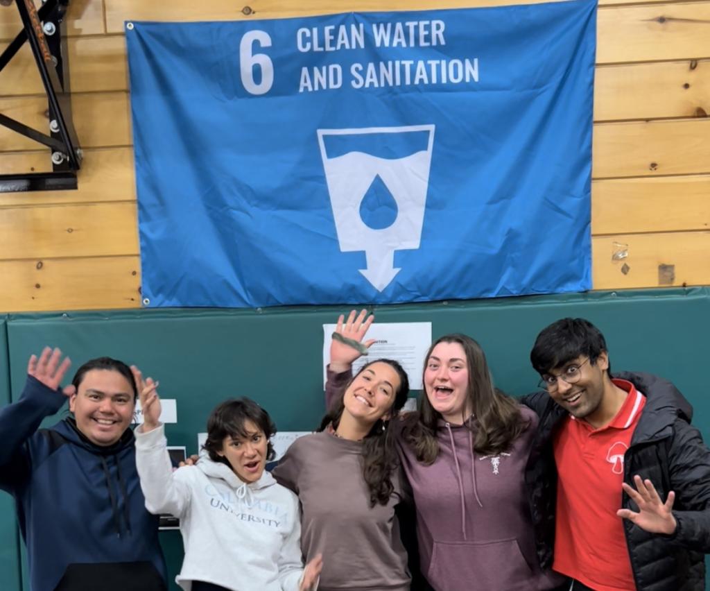 Adrienne and her team at CAMP stand in a gymnasium smiling and waving. They're standing in front of a sign with their UN Sustainable Development Goal - about sanitation and water