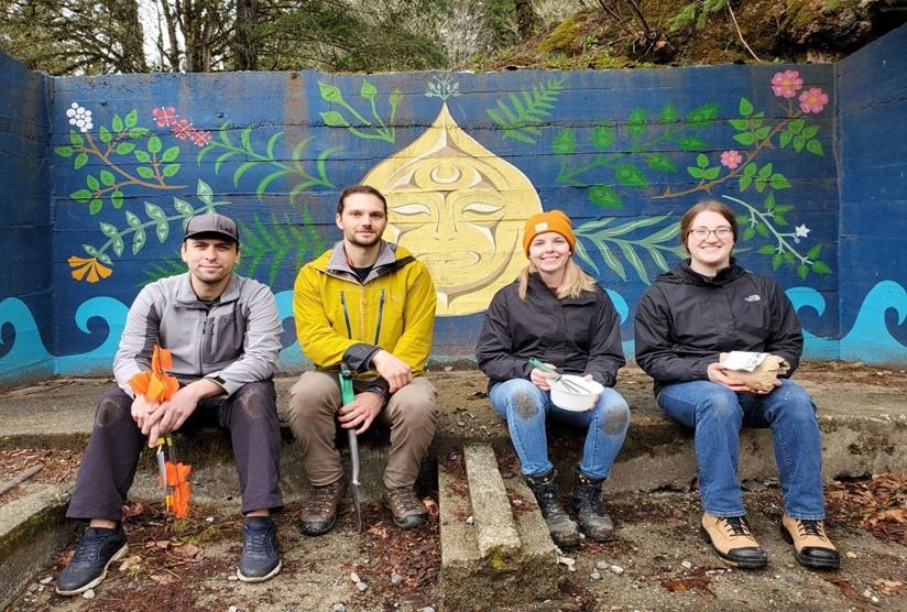Members of TerraMend Consulting Ltd. sitting in front of a mural painted over concrete infrastructure at one of three remediation plot sites (SṈIDȻEȽ, near Tod Inlet).