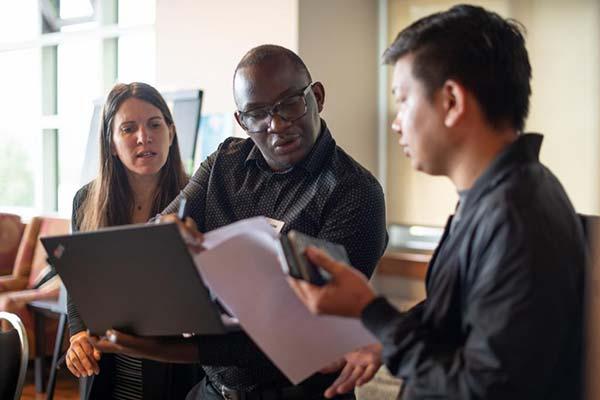 Three people seated in a row look toward one person's notes attentively.