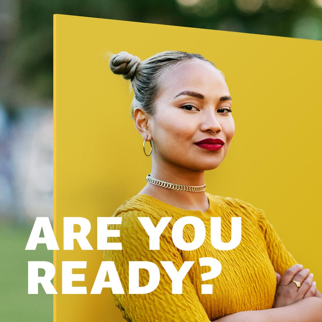 A person in yellow with red lipstick and spacebuns stands cross-armed in front of a yellow background.