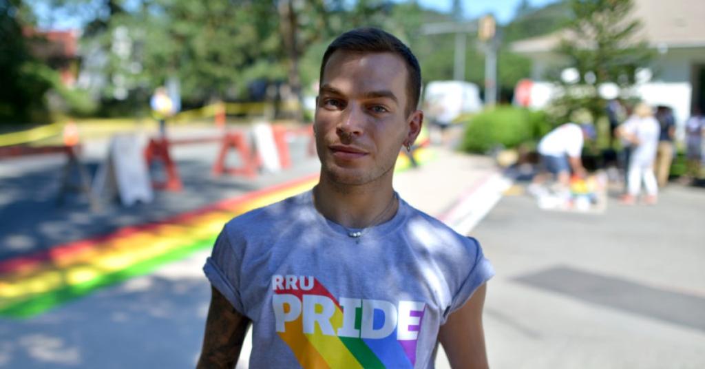 A student in an RRU Pride t-shirt stands in front of an rainbow crosswalk being painted on campus.