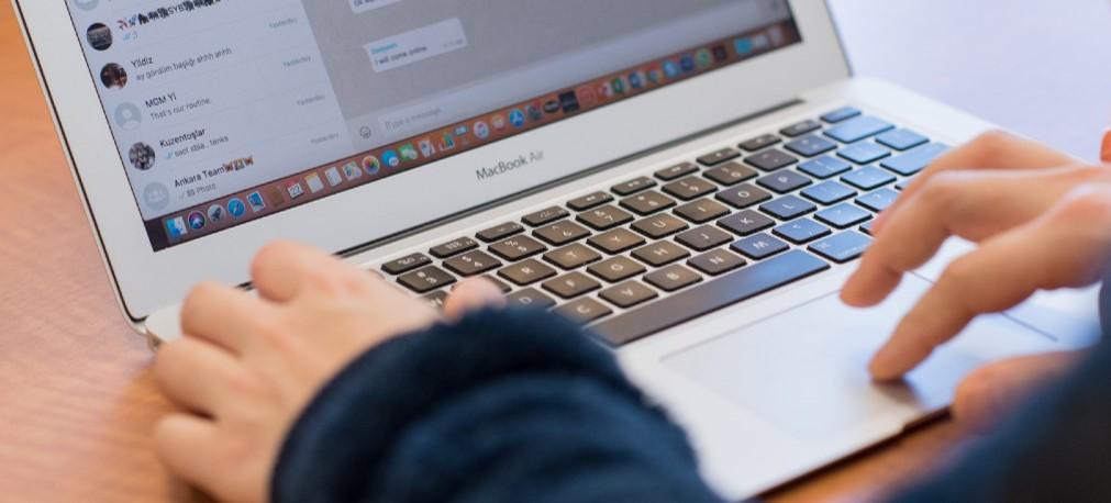 A silver laptop, opened to a social media page, is viewed from over someone's shoulder.  The person's hands and black sleeves are visible over the keyboard.