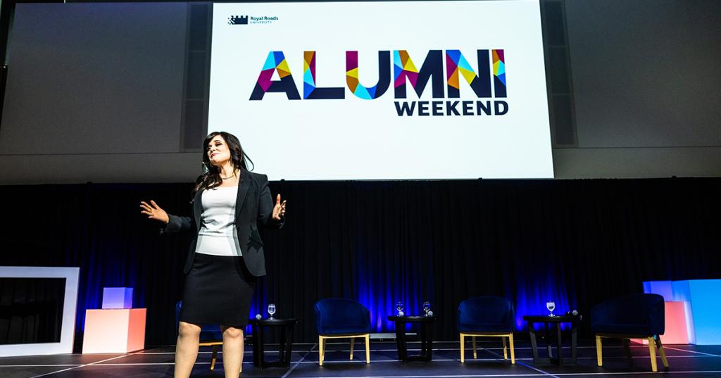 An alumni giving a bold presentation on stage with a big screen behind her that says "Alumni Weekend" in RRU colours.