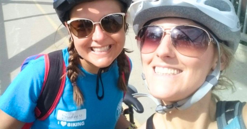 Two people pose for a selfie while smiling and wearing bike helmets and sunglasses