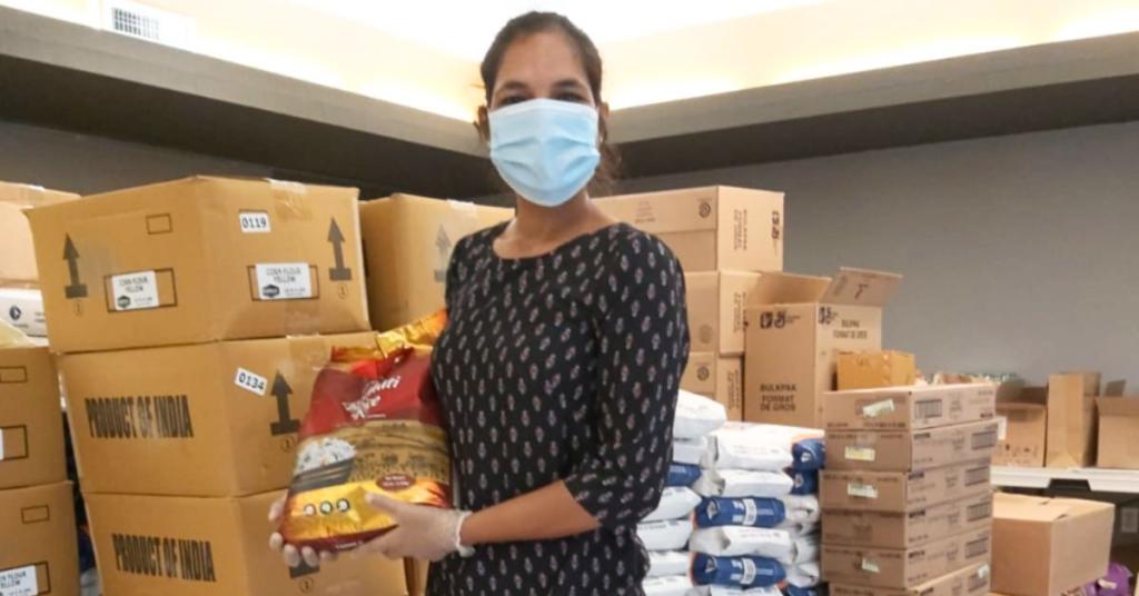 Narinder Kaur stands in a food storeroom holding a bag of food while wearing a mask