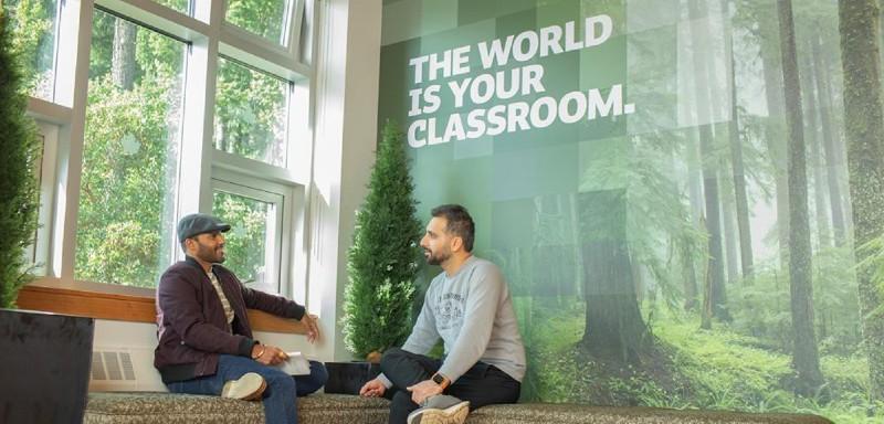 Two Royal Roads University students sit in the foyer of a building, next to a bright window and a backdrop that reads "The world is your classroom".