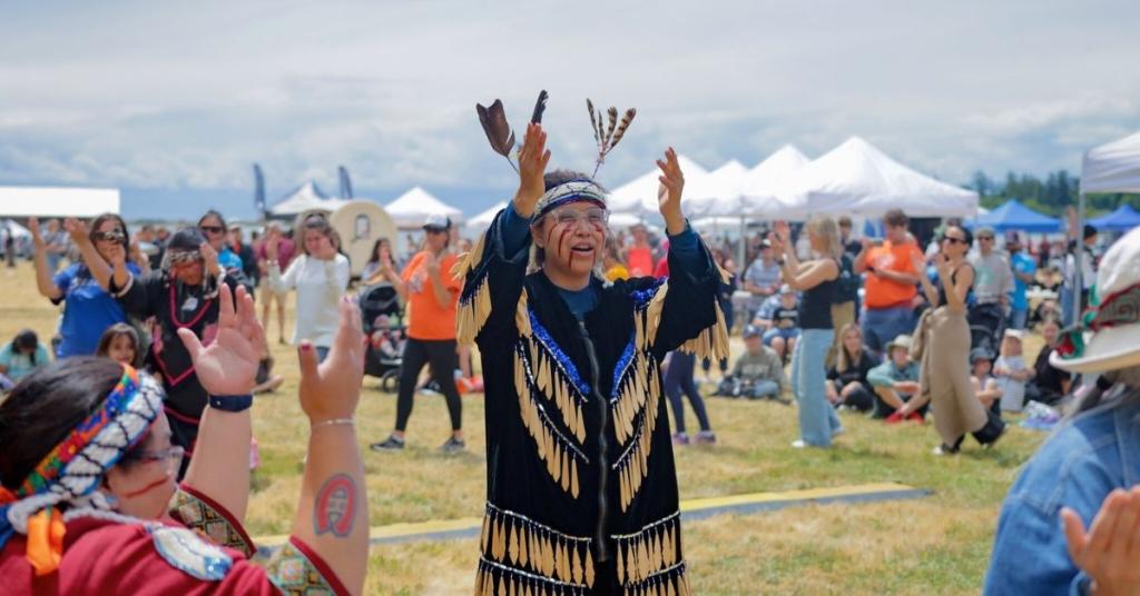 A performer raises hands in the air with dancers gathered around at the 2025 National Indigenous Peoples Day celebration at Royal Roads University