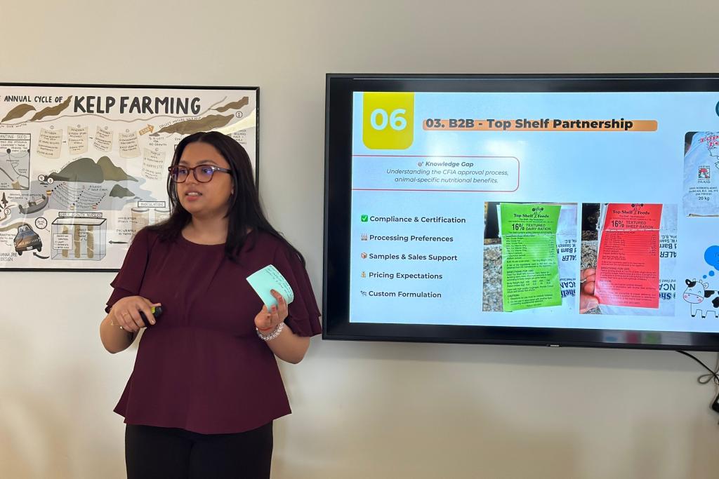 A woman gestures during a presentation, a TV screen can be seen behind her.