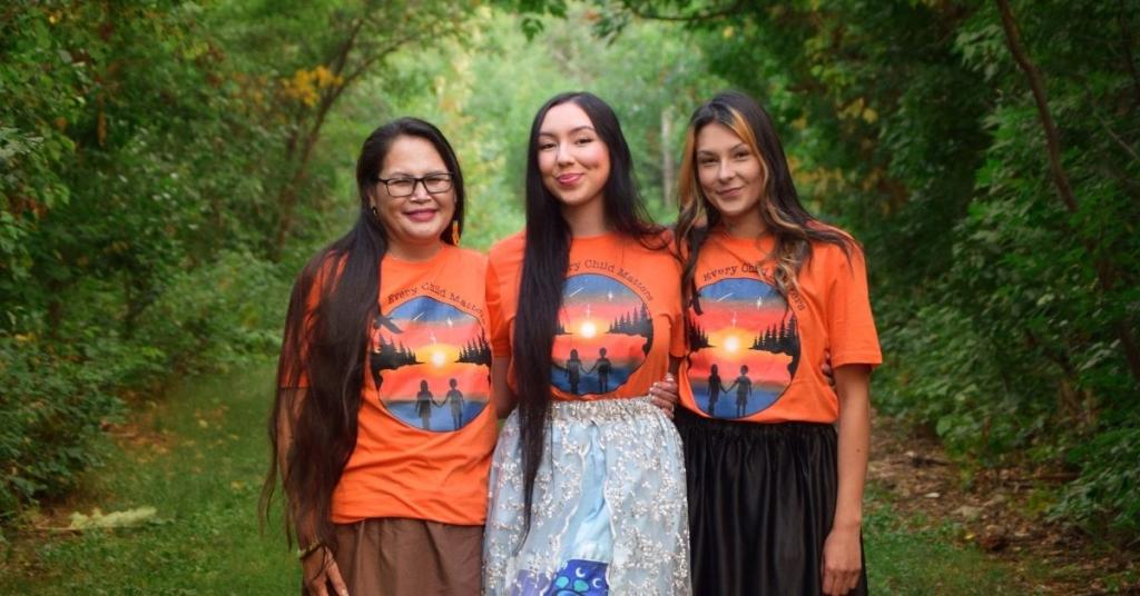 Three women wear orange t-shirts and ribbon skirts with a forested background.
