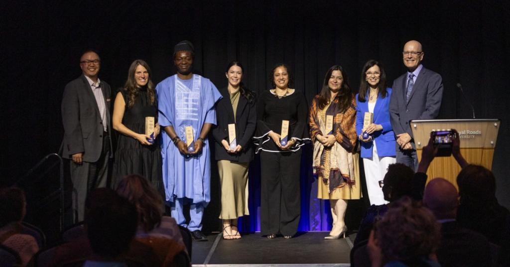 The 2025 winners of the Alumni Changemaking Achievement Awards stand on the stage in the Dogwood Auditorium holding their awards, beside Chancellor Nelson Chan and President Philip Steenkamp.