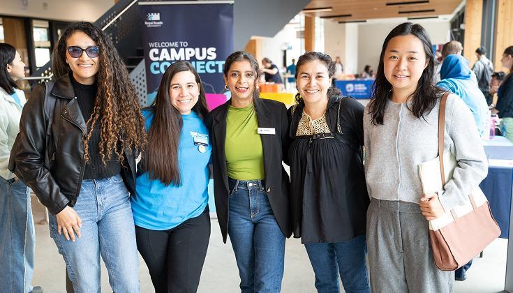 A group of five RRU students smiling and standing together at a Campus Connect event indoors, with tables and event signage visible in the background.