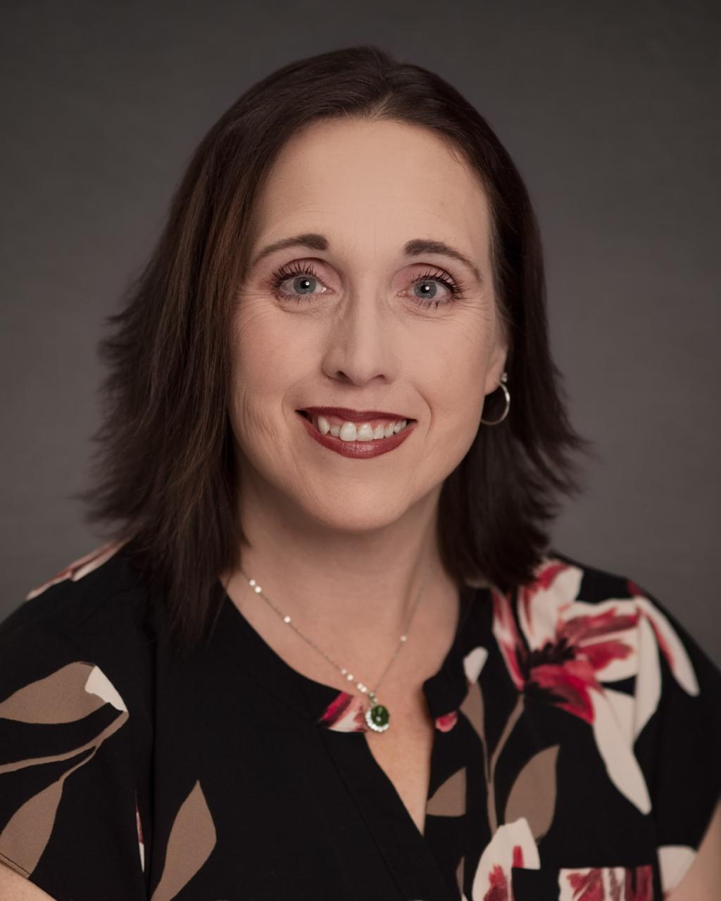 woman with dark hair that just touches her shoulders smiling wearing a black, brown, white and red floral blouse