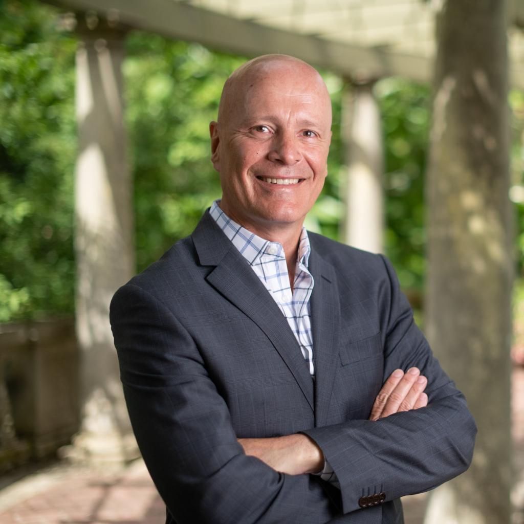 President Philip Steenkamp in the Italian Garden with a suit and no tie. His arms are lightly crossed and he's smiling. 