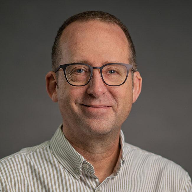 man with close cropped brown hair wearing glasses and a white collared shirt with black pinstripes. He's grinning slightly.