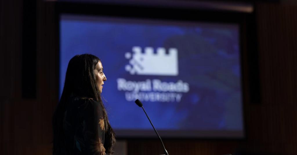 Emily Mulroney standing in profile at a podium with a screen displaying the Royal Roads University logo beside her.