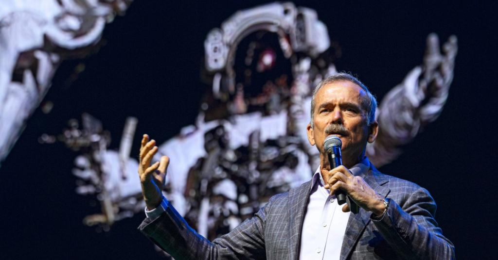Chris Hadfield presenting into a microphone, with a picture of an astronaut in the background