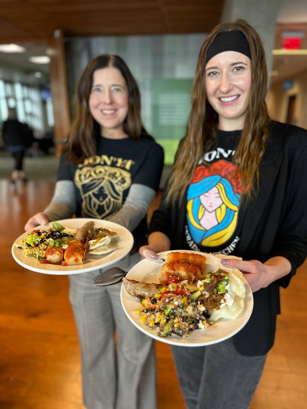 Two women hold plates of food including sausages, cabbage rolls and perogies.
