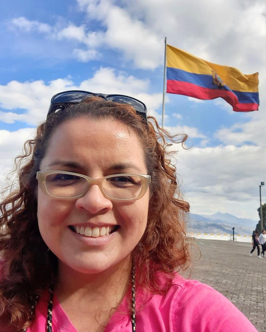 A woman smiles while taking a selfie. An Ecuadorian flag can be seen flying in the background amid a blue sky with white clouds.