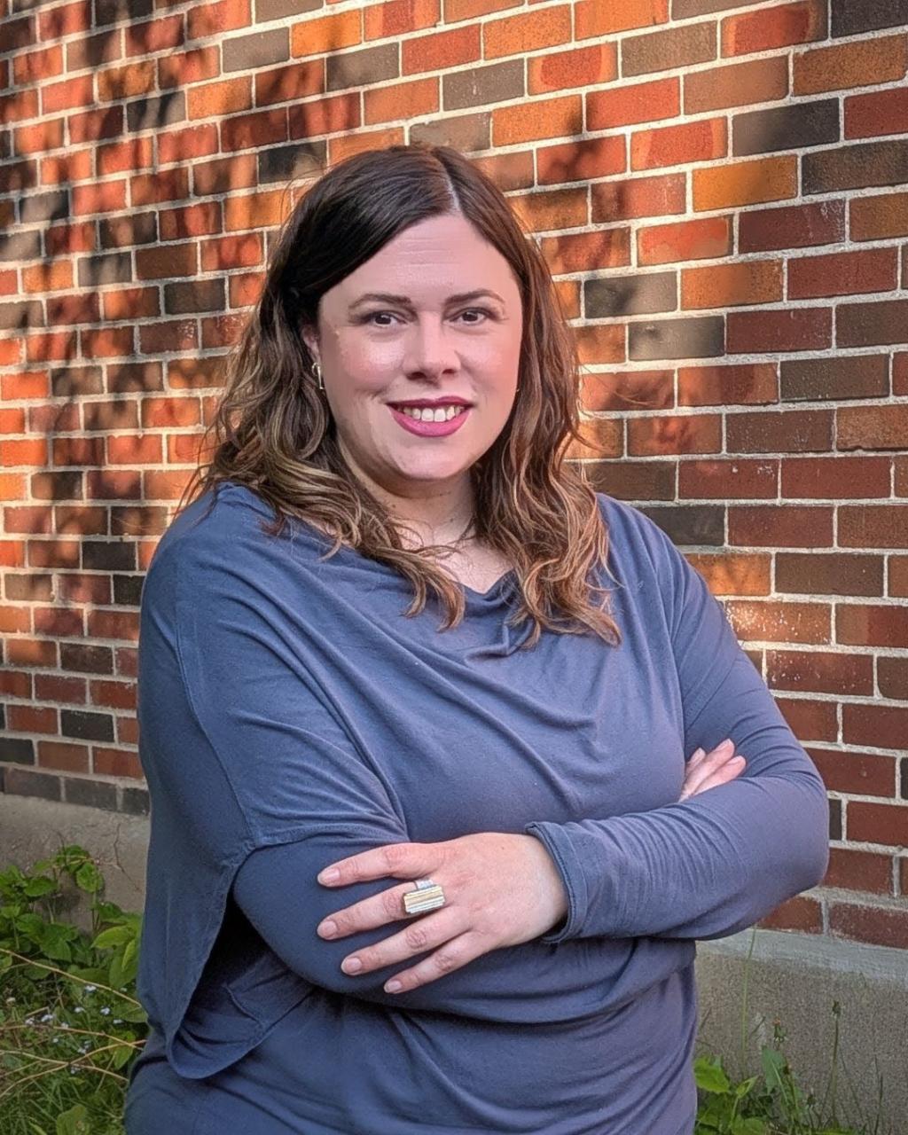 A woman smiles with crossed arms in front of a brick wall.