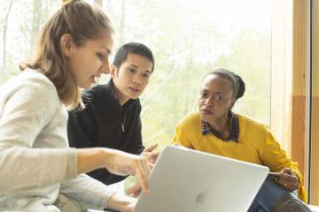 Group of people looking at a laptop screen with trees visible through window behind them.