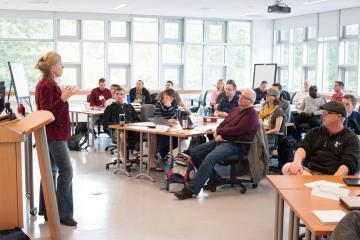 Instructor standing at front of classrom speaking to students.