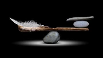 A piece of wood balanced on a rock with a feather on one side and rocks on the other and they're balanced