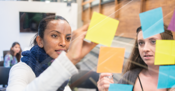 Individuals writing on and organizing colourful sticky notes on clear display board.