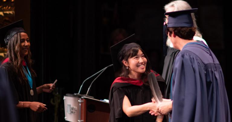 Graduate wearing academic regalia shakes hands on stage at Convocation.