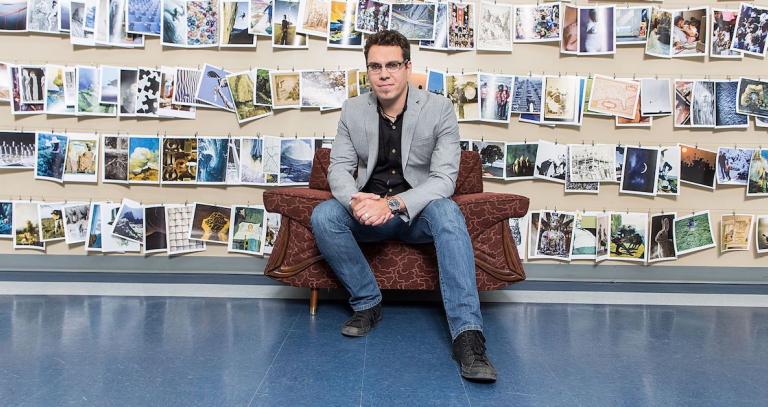 MA in Leadership graduate sits in a chair in front a wall covered in photographs.