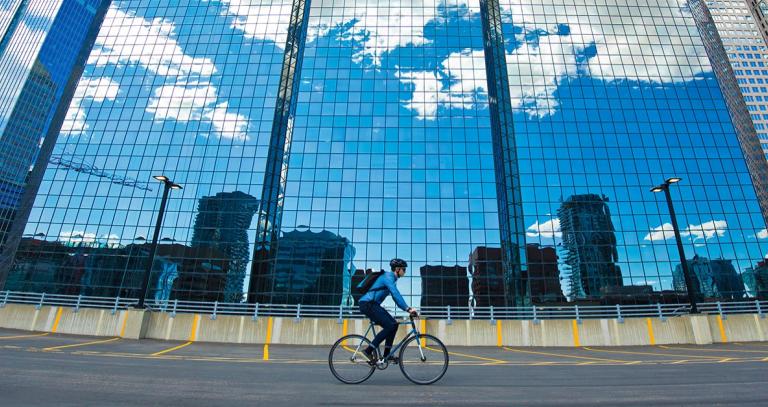 Person riding their bike on a highway next to a glass building reflecting a blue sky