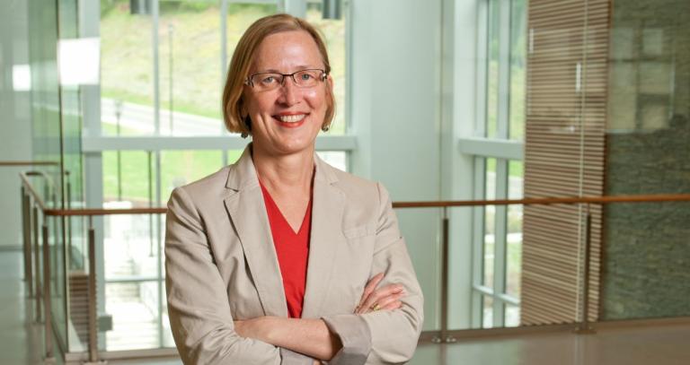Smiling woman in red shirt and tan blazer against a modern building interior background