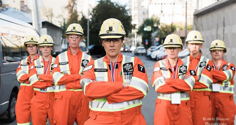 Group of seven women wearing hard hats and orange safety jumpsuits stand with their arms crossed -- they are a mine rescue team.